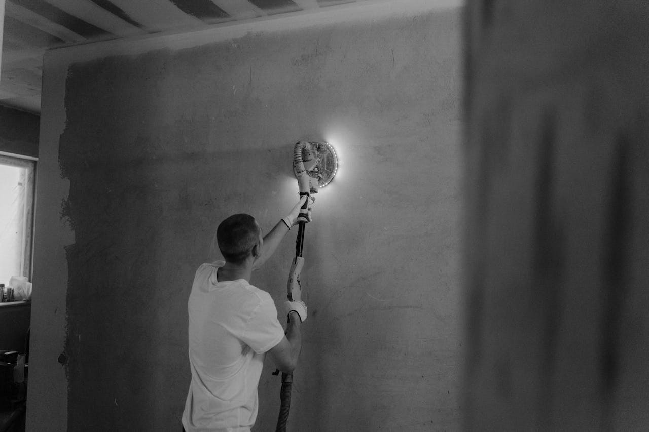 A man using a power sander to renovate an interior wall in a monochrome setting.